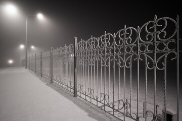 Night winter view of the fence. Street lamps illuminate a metal fence covered with frost. Snow covered sidewalk. Cold winter weather. Dark street.