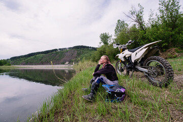 Beautiful female biker wearing pink dreadlocks sitting along river near to her enduro offroad motorcycle