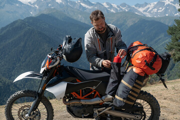 Man motorcyclist traveler standing next to dirt motorcycle and packing hiking bags with amazing mountains landscape on background