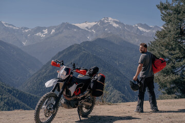 Male motorcyclist traveler standing in mountains near his motorcycle with large hermetic bag on his shoulder. Moto trip