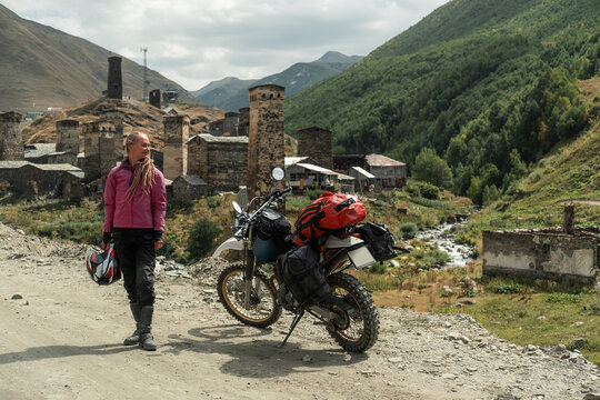 Woman traveling solo on motorcycle off-road in mountains, standing near stone old village admiring the scenery