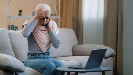 Obraz premium Muslim islamic arabian woman in pink hijab sitting on sofa working using laptop feel headache stress migraine severe pain tired sad female freelancer having health troubles high pressure overtime work
