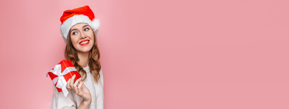 Young caucasian woman with santa claus hat and gift box smiling isolated over pink background. web banner. copy space