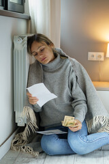 Sad woman covered with grey blanket sits on floor and looks at bills with huge communal payments....