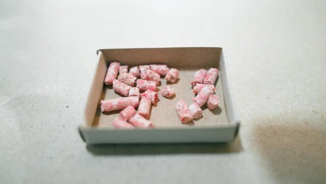 Pink granules of poison from rodents are poured into a cardboard box close-up. Flavored, granular, brightly colored mouse poison. Fighting rats with chemicals that affect blood clotting