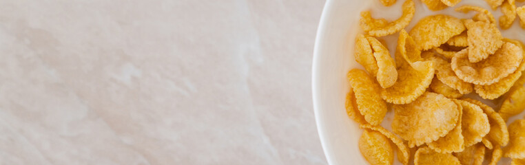top view of crispy corn flakes in bowl with spoon and milk on marble surface, banner.