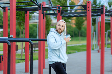 Obraz premium A man with a braid hairstyle in the early morning drinks coffee on the sports ground.