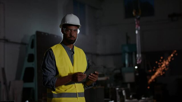 Potrait of young heavy industry worker wearing safety glasses and helmet, using tablet and smiling at camera.