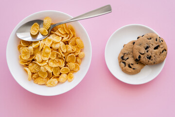 top view of bowl with tasty corn flakes and spoon near saucer with chocolate chip cookies on pink.