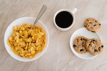 top view of bowl with corn flakes near cup of coffee and chocolate chip cookies on marble surface.