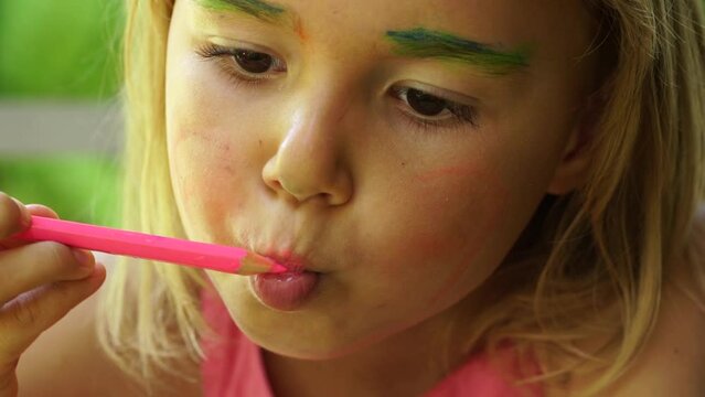 Close-up of little cute girl doing makeup with color pencils. She paints her face imitating mom or an adult woman