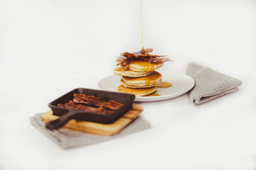 Maple Syrup being poured on to pancake stack topped with crispy bacon.  Cast-iron skillet in the foreground.
