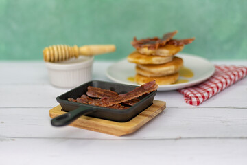 Mini cast-iron skillet with crispy bacon and pancake stack with  honey topping in the background