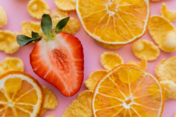 close up view of fresh sliced strawberry around crispy corn flakes and dried oranges on pink.
