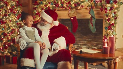 Slowmo of bearded Santa Claus in red costume talking to cute little African American girl sitting on his laps with beautifully decorated Christmas tree and fireplace in background