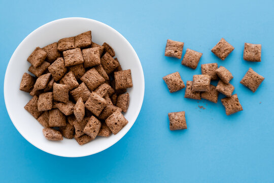 Top View Of Chocolate Cereal Puffs And White Bowl Isolated On Blue.