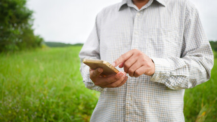 A farmer is using a mobile phone standing in a field against a green field backdrop.