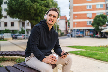 Young guys listening to music while texting and talking on his phone and drinking coffee in his hoodie outside during the day