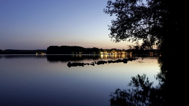 Time lapse shot of some rocks in the water of small lake Brombach (kleiner Brombachsee) and trees while a colorful dusk with some stars appearing