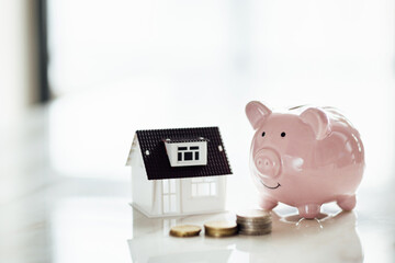 Pink piggy bank with tiny house on white marble background and blurred bokeh with copy space....