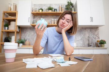 Unhappy and depressed woman sitting in the kitchen at the table, holding piggy bank and looking at...