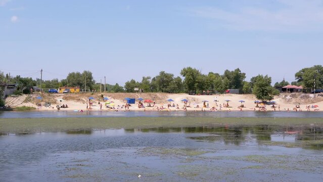 Panoramic View of City Beach with Umbrellas, People Resting on the River Bank. Coastline, blooming green river, beach area. Blue sky, white sand, bathing tourists. Nature, summer landscape. Weekend.