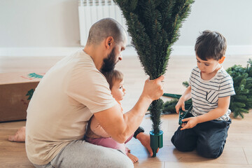 Father with his children assembling the Christmas tree in their living room preparing for the winter holidays. Christmas, holiday, winter concept. Happy family. - obrazy, fototapety, plakaty
