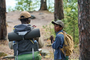 Cute boy with backpack looking at his grandfather while talking to him during travel in pine tree forest on summer weekend