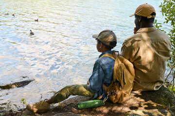 Naklejka premium Back view of youthful African American boy with rucksack and his grandfather sitting in front of lake or river during halt