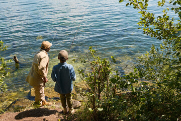 Rear view of youthful boy and his grandfather with rods standing in front of lake on sunny summer...