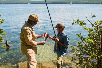 Side view of happy mature black man in activewear showing fish to his grandson with rod while both standing against waterside - Powered by Adobe