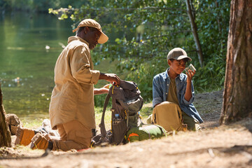 Aged African American man unpacking his rucksack while standing on knees on footpath in front of his grandson with jar of grasshoppers