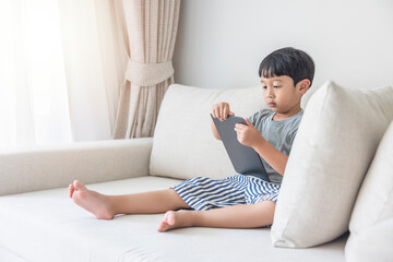 Adorable happy Asian boy wearing a gray shirt and blue-white striped shorts is having fun playing with his tablet on a cream sofa. looking at the mobile screen