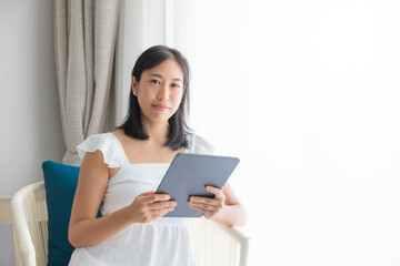 Adorable happy Asian woman wearing white dress is having fun playing with his tablet on a cream sofa. looking at camera