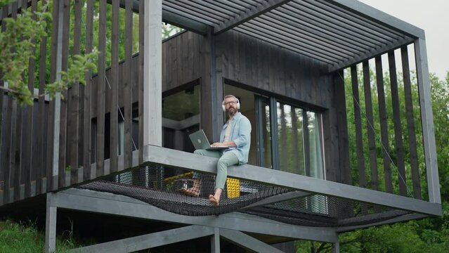 Man sitting on tiny house terrace working on laptop, listening to music