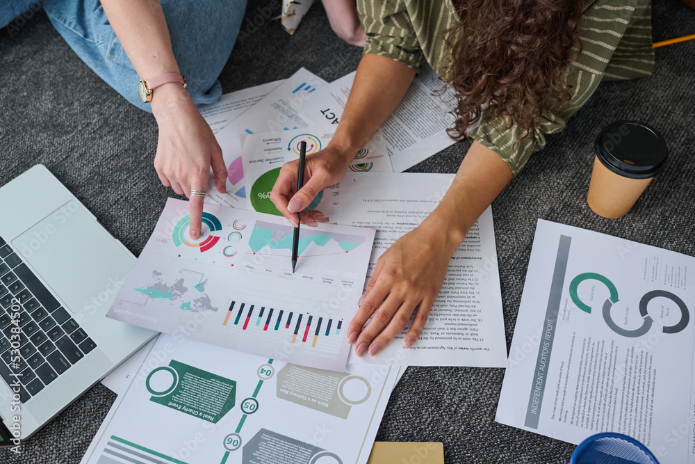 Poster above view of hands of two young businesswomen discussing documents with graphic financial data on t
