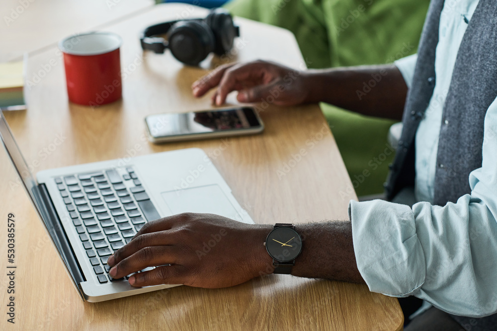 Poster hands of young black man in formalwear keeping hand over laptop keypad while scrolling in smartphone