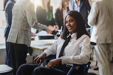 Portrait of a young African American businesswoman in a wheelchair smiling in the office - group of multi-ethnic businesspeople in the background - business lifestyle concept - obrazy, fototapety, plakaty