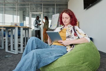 Young serious employee in casual attire sitting in armchair and networking or preparing for presentation in office