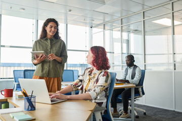 Happy young office worker with digital tablet communicating with female colleague sitting by workplace in front of laptop during talk