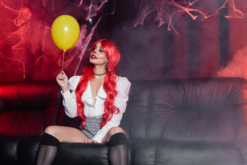 young redhead woman in clown makeup looking at yellow balloon on dark background with fog and spiderweb.
