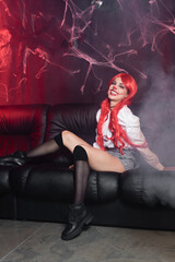 Obraz na płótnie Canvas excited redhead woman in clown makeup and black knee socks sitting on leather couch near spiderweb on dark background.