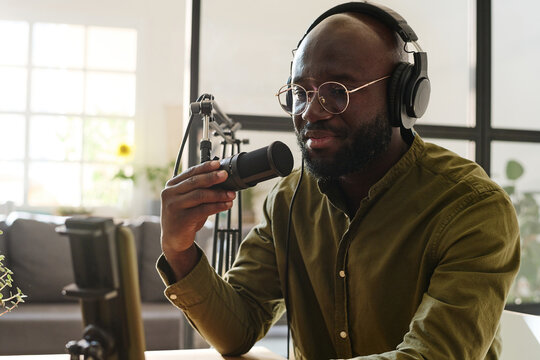 Young black man in eyeglasses and headphones looking in smartphone camera during livestream or recording new video for online audience