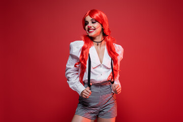 redhead woman with clown makeup posing in white blouse and shorts with suspenders isolated on red.