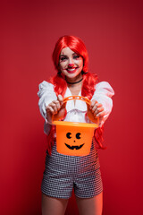 cheerful woman in clown makeup and bright wig showing halloween bucket isolated on red.