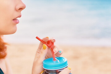Close-up of a woman's lips and an alcoholic cocktail with a straw in her hands. Sea beach in the background. Lady on vacation in the tropics.