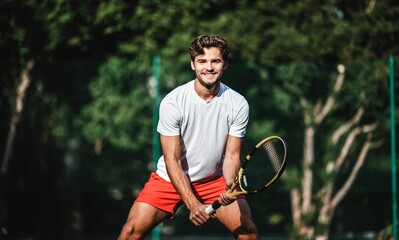 A handsome Caucasian tennis player smiling, carrying his racket and ready to play on an outdoor court, with trees in the background. Concept of joy of playing sports and recreation outdoors.