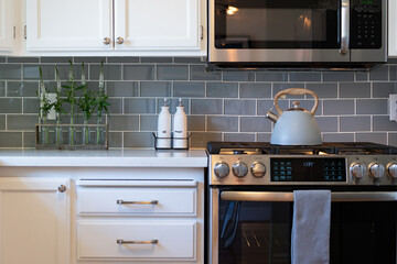 Modern kitchen details of white counter and cabinets, gas stove with teapot, and gray tile backsplash.
