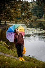 portrait of a beautiful smiling woman in brown poncho, beautiful bracelets, earrings in yellow rubber boots, backpack and with rainbow umbrella walking near lake with wild ducks in autumn park