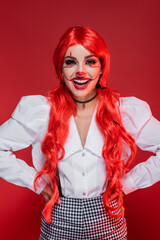 smiling woman with clown makeup and bright hair standing with hands on hips isolated on red.